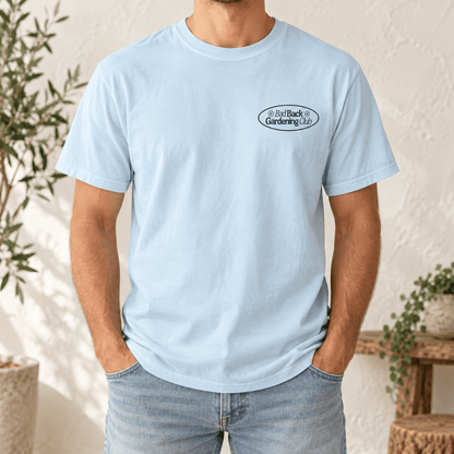 A man wearing a light blue crewneck t-shirt with a small black oval logo on the left chest that reads 'Bad Back Gardening Club'. He is also wearing light-wash blue jeans, standing in a brightly lit room with a textured wall and potted plants.