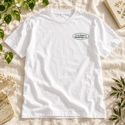 A white short-sleeve crewneck t-shirt with a small green and blue 'Bad Back Gardening Club' logo on the left chest, styled flat on a beige linen fabric with green leaves and dried flowers.