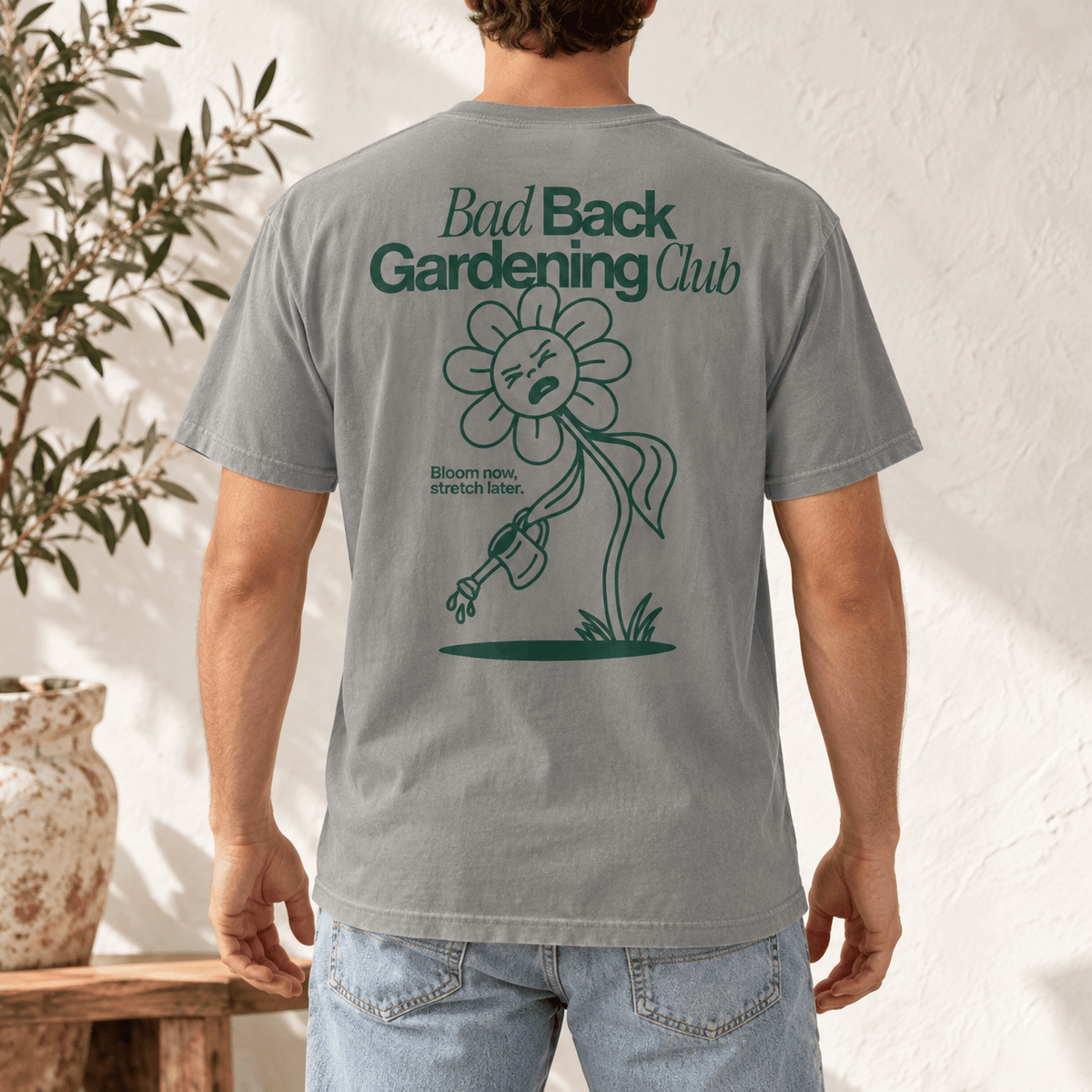 A man wearing a gray t-shirt with a green graphic on the back that reads 'Bad Back Gardening Club' and shows light blue jeans, standing in front of a textured white wall with a potted plant on the left.