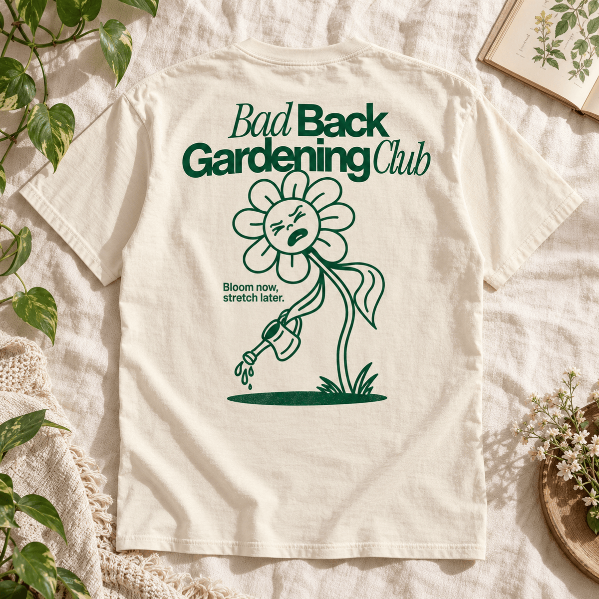 An off-white short-sleeve T-shirt laid flat on a textured cream fabric, surrounded by decorative plants, flowers, and a book. The back of the shirt features a dark green graphic of a sad flower watering plants, with the text 'Bad Back Gardening Club' and 'Bloom now stretch later.