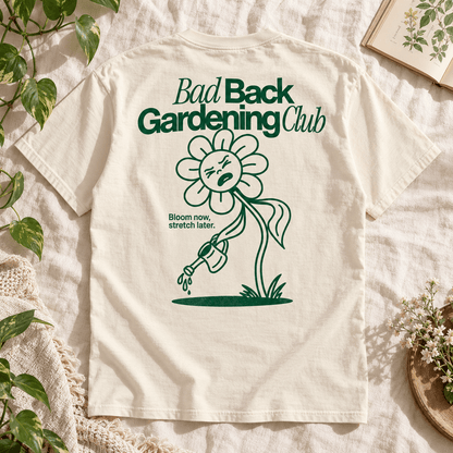 An off-white short-sleeve T-shirt laid flat on a textured cream fabric, surrounded by decorative plants, flowers, and a book. The back of the shirt features a dark green graphic of a sad flower watering plants, with the text 'Bad Back Gardening Club' and 'Bloom now stretch later.