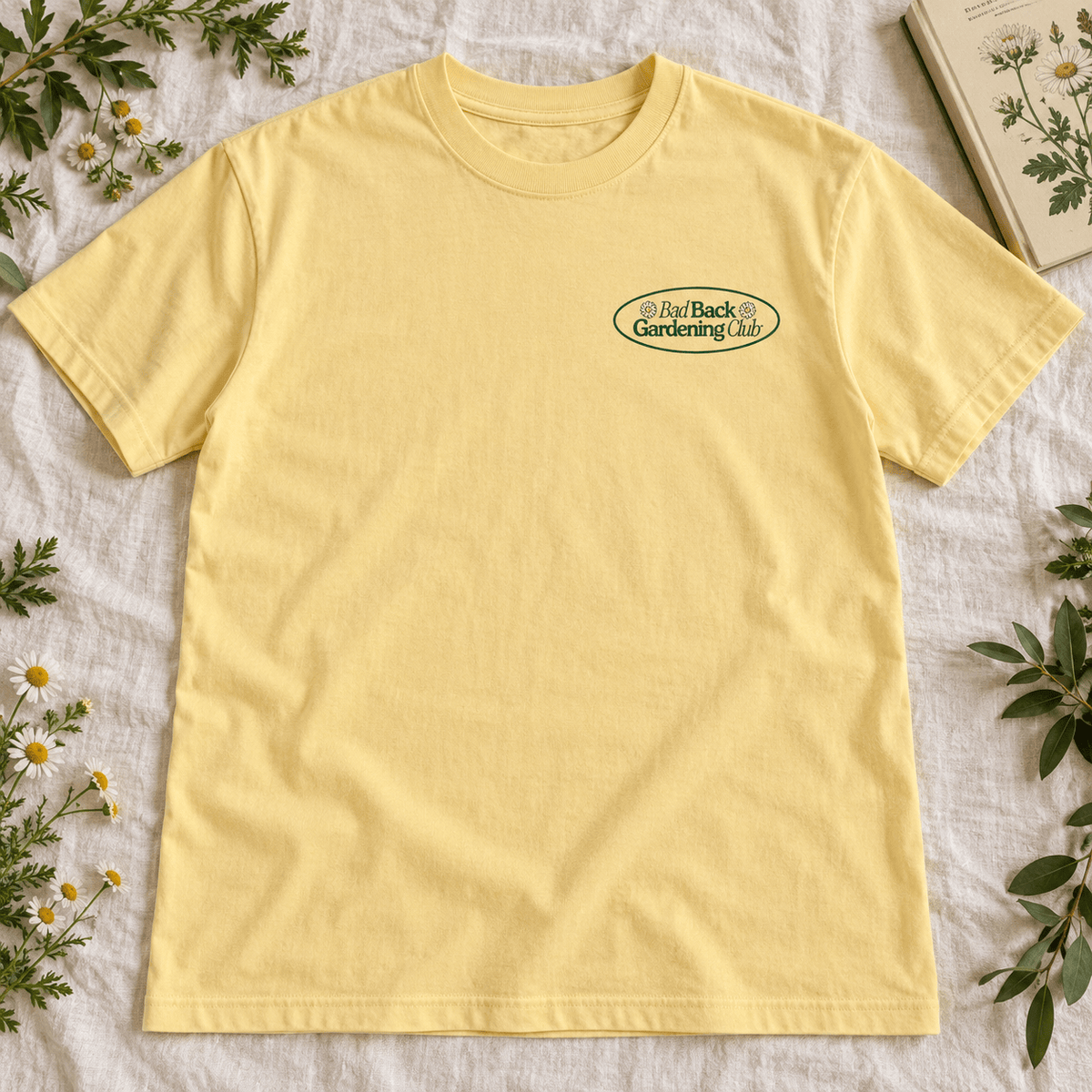 A pale yellow short-sleeve crewneck t-shirt with an oval 'Bad Back Gardening Co.' logo on the left chest, styled flat on a textured white fabric with green leaves and small white flowers.