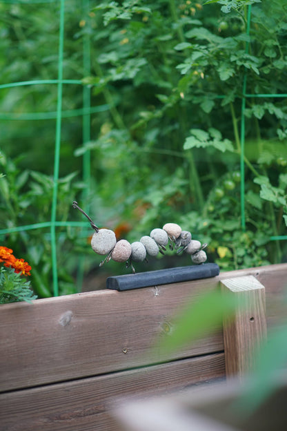 Stone caterpillar garden sculpture made from river rocks and wire, mounted on a dark base, displayed on a wooden raised garden bed