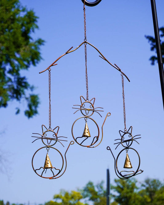 Three rusty wire cat-shaped wind chimes with brass cone bells hanging outdoors against a blue sky