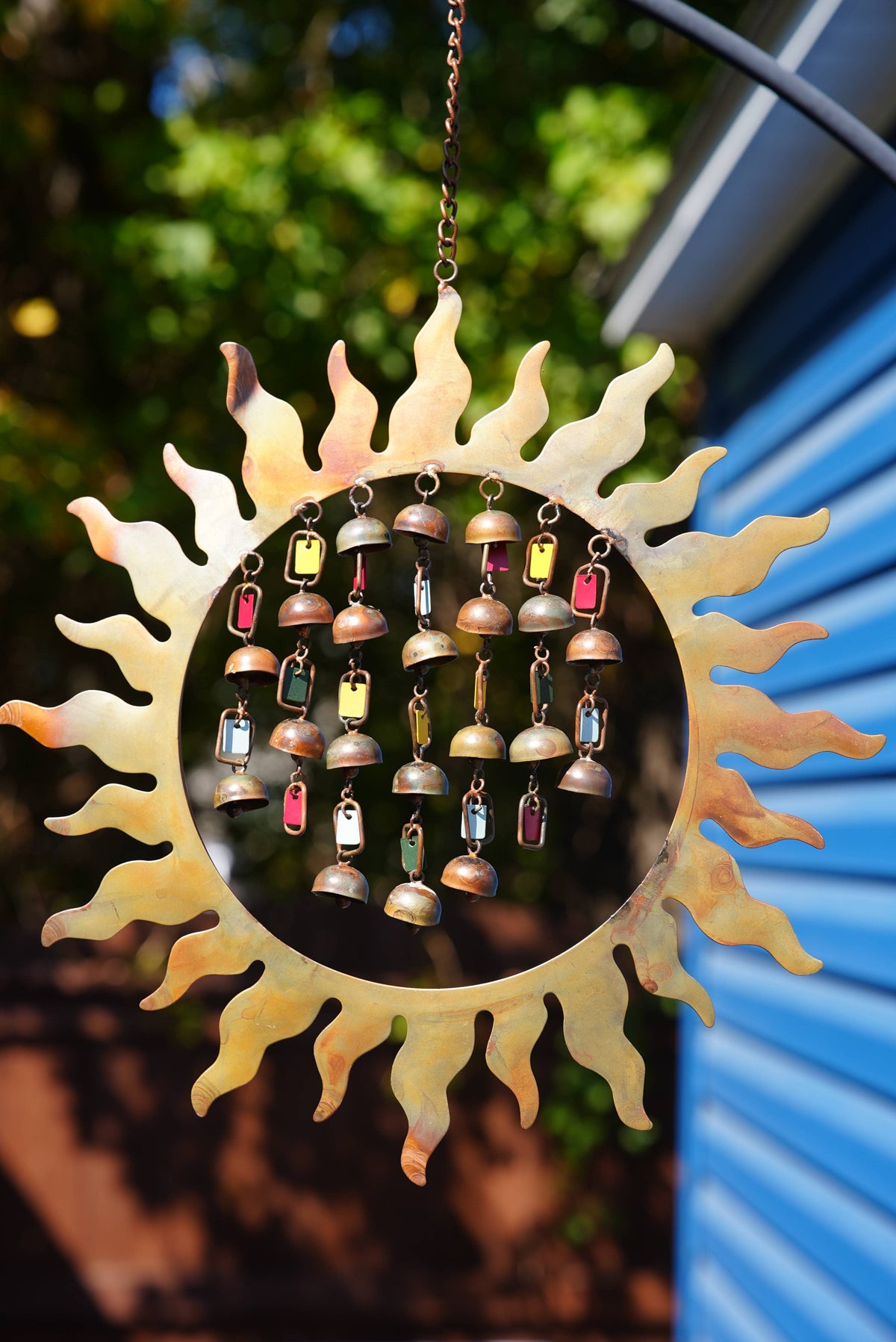 A sunburst metal wind chime with a circular center containing small hanging bells and colorful rectangular charms, photographed outdoors against a blurred background of green foliage and a blue corrugated wall.