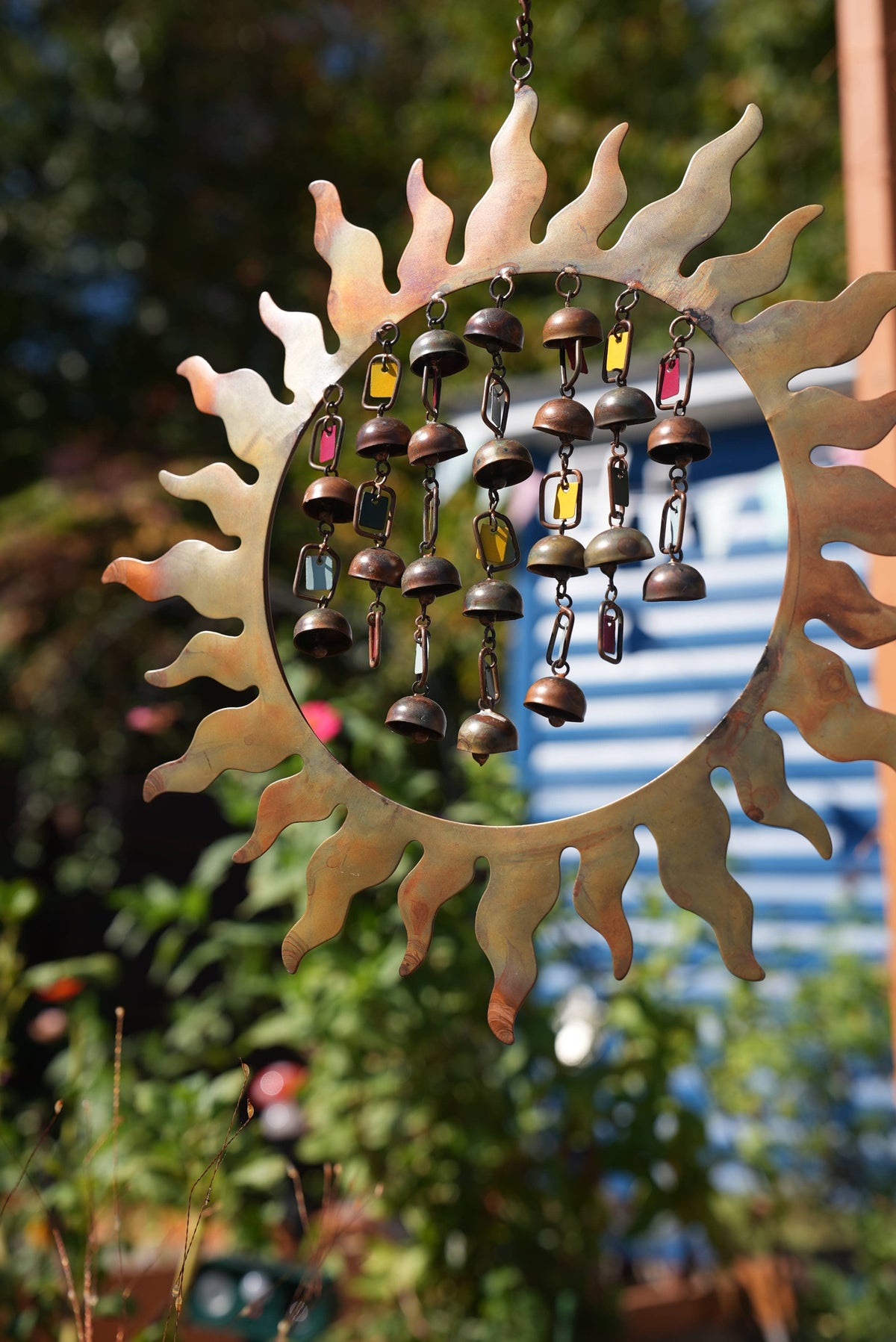 A metal sun-shaped hanging garden ornament with a wind chime inside, featuring bronze-colored bells and colorful rectangular charms, set against a blurred outdoor garden background.