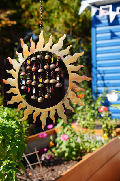 A sun-shaped metal wind chime hanging in a garden. The center features a circular black section filled with small hanging bells in various colors, surrounded by a gold-toned sunburst frame.