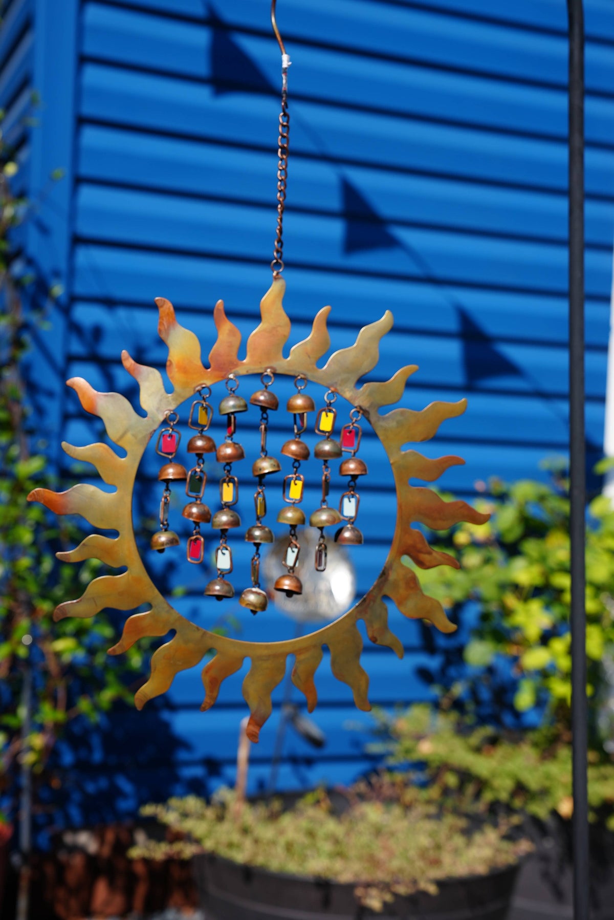 A decorative metal sun ornament hanging outdoors in front of a blue house wall, with small wooden bells and colorful hanging accents in the center.