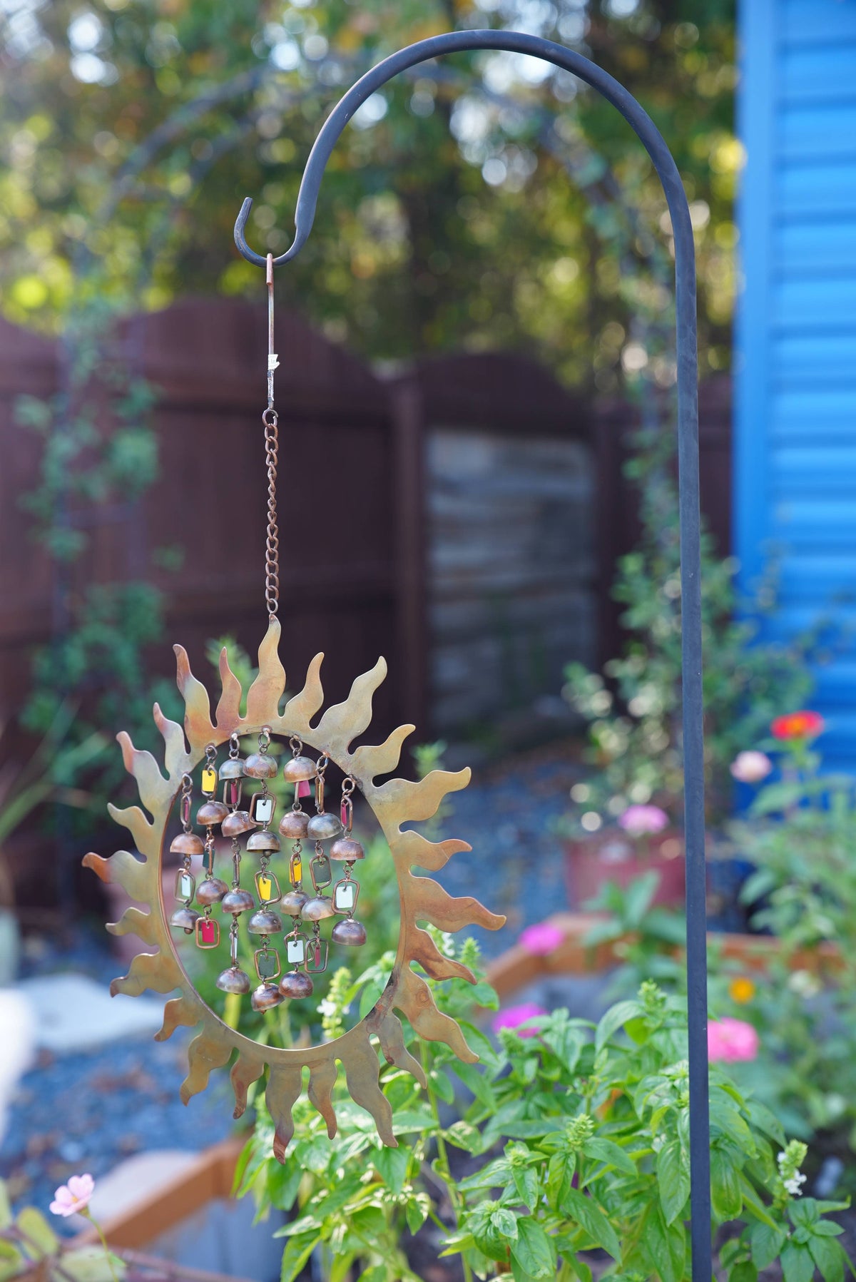 A metal sunburst wind chime with hanging glass beads is displayed on a black metal garden hook in a garden setting.