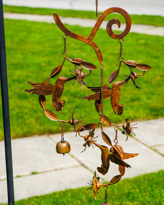 Copper-toned metal wind chime featuring hummingbirds, flowers, and small bells hanging from a decorative scrolled frame