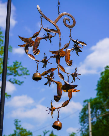 Copper-toned metal wind chime with hummingbird and flower figures, hanging chains, and two round bells against a blue sky