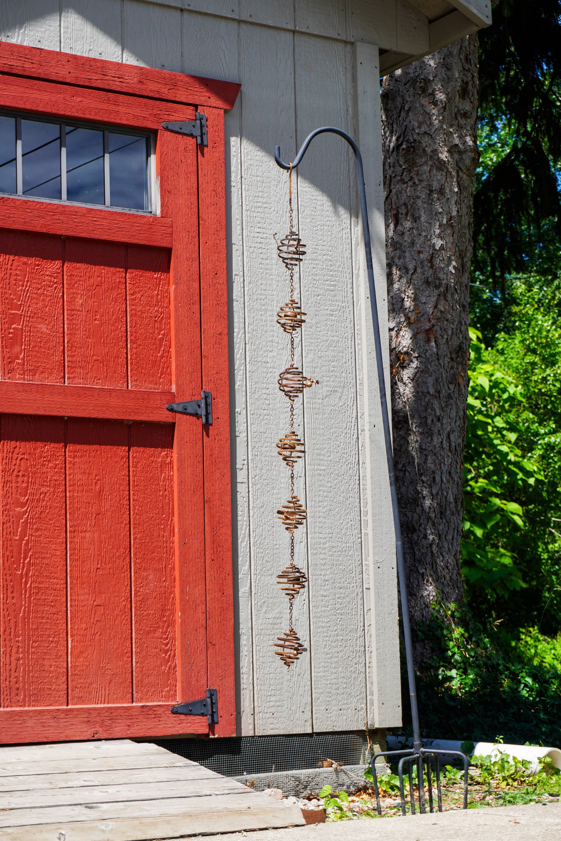 Rustic metal spiral bee ornaments linked by chain, hanging from a shepherd's hook beside a red barn door