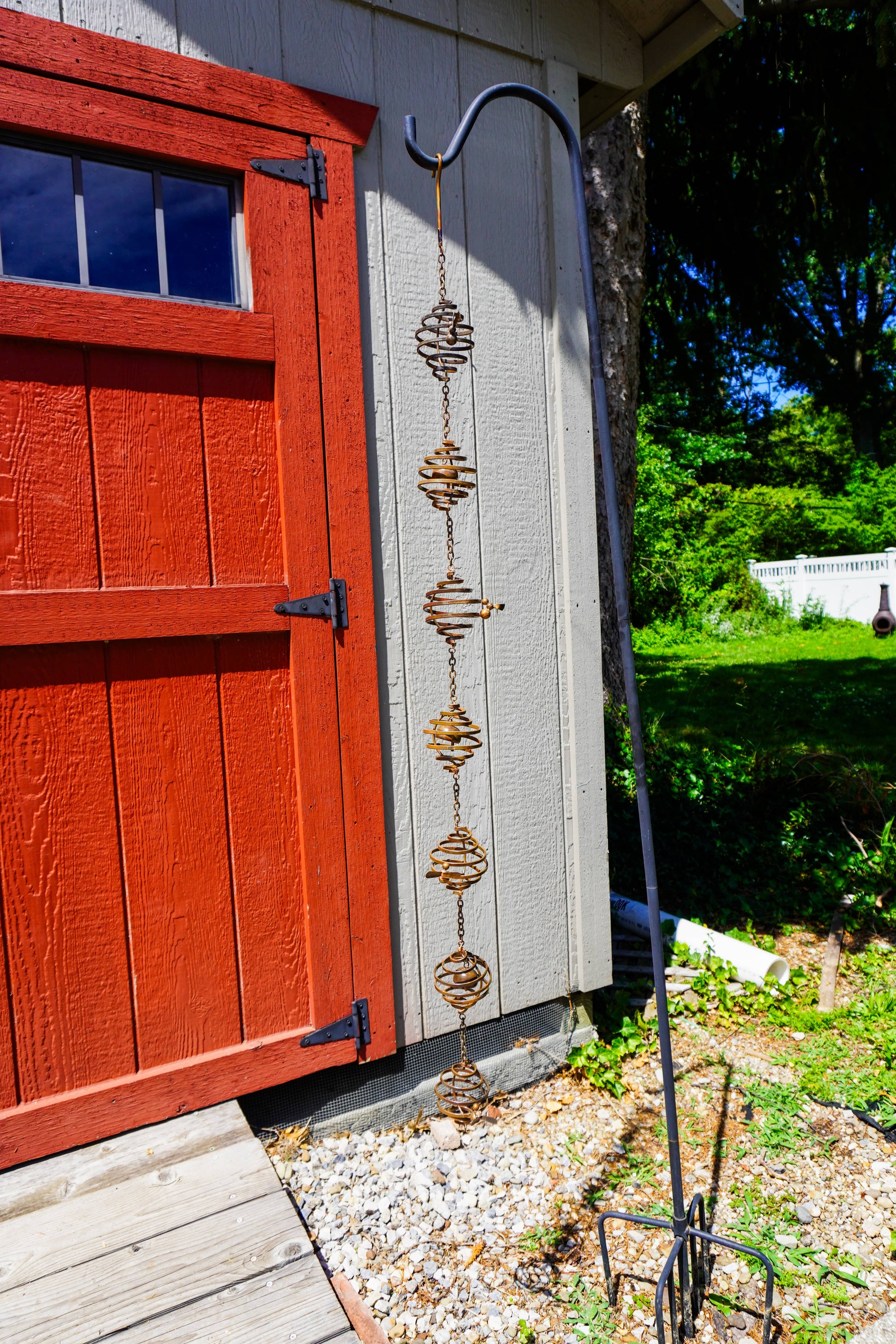 Rusty metal spiral coil ornaments linked by chains hanging from a shepherd's hook beside a red barn door