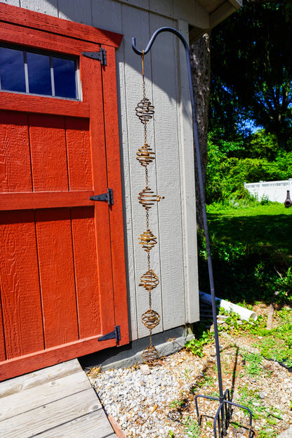 Rusty metal spiral coil ornaments linked by chains hanging from a shepherd's hook beside a red barn door