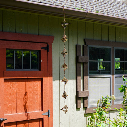 Spiral metal wind spinner with stacked coiled orbs hanging from a hook on a green wooden house exterior