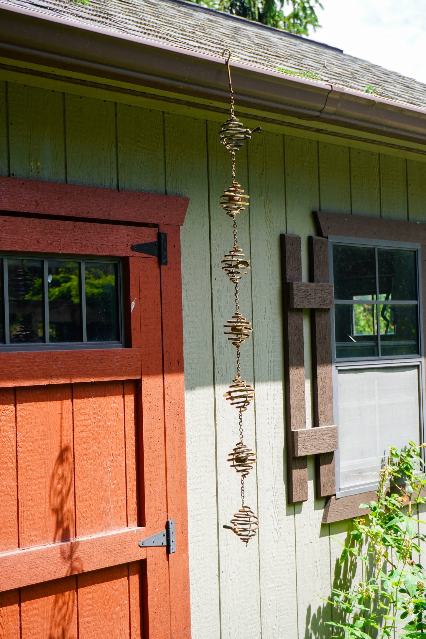 Hanging metal spiral wind spinner with six stacked coil orbs linked by chains, mounted on a green wooden building exterior