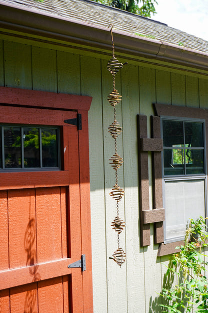 Hanging metal spiral wind spinner with six stacked coil orbs linked by chains, mounted on a green wooden building exterior