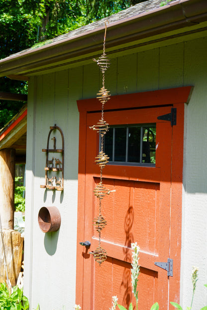 Brass spiral wind spinner hanging from eave of red and green garden shed with decorative metal ornaments nearby