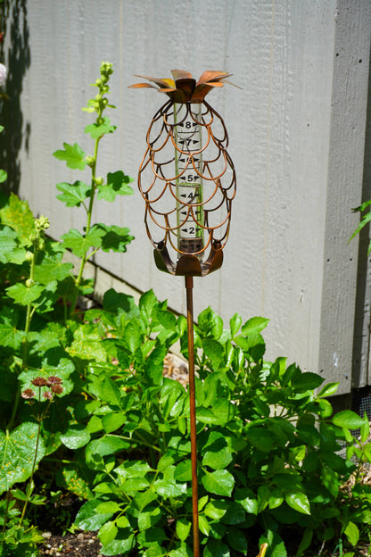 Copper-toned metal pineapple-shaped rain gauge with glass tube on a garden stake surrounded by green plants