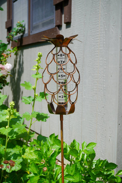 Copper metal pineapple-shaped rain gauge garden stake with glass tube marked 2–8 inches, surrounded by green garden plants