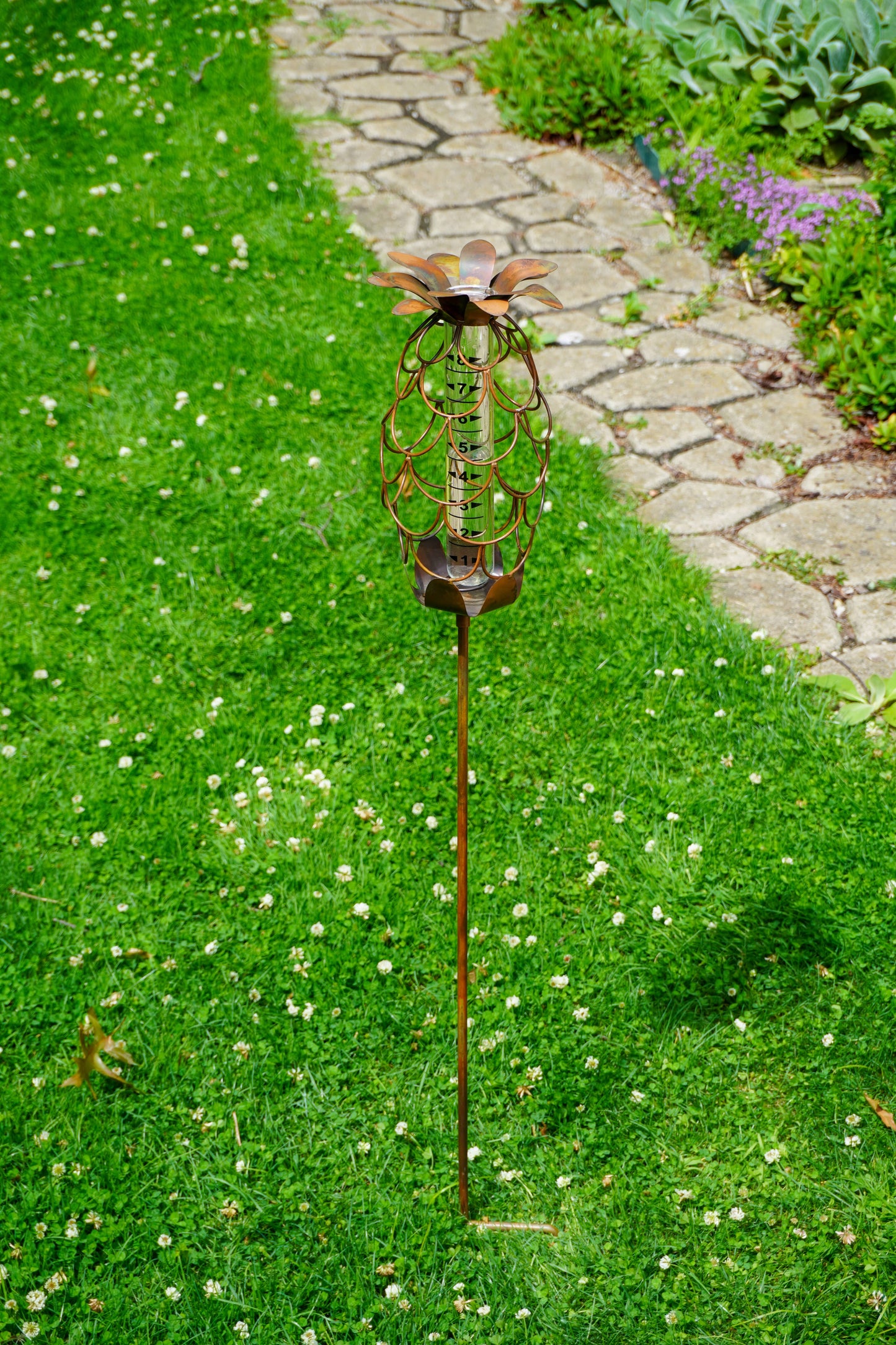 Decorative copper-toned metal pineapple rain gauge on a tall stake, displayed in a green garden lawn