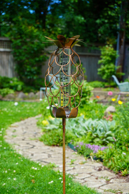 Copper-toned metal pineapple-shaped rain gauge garden stake with glass tube insert, displayed outdoors in a green garden