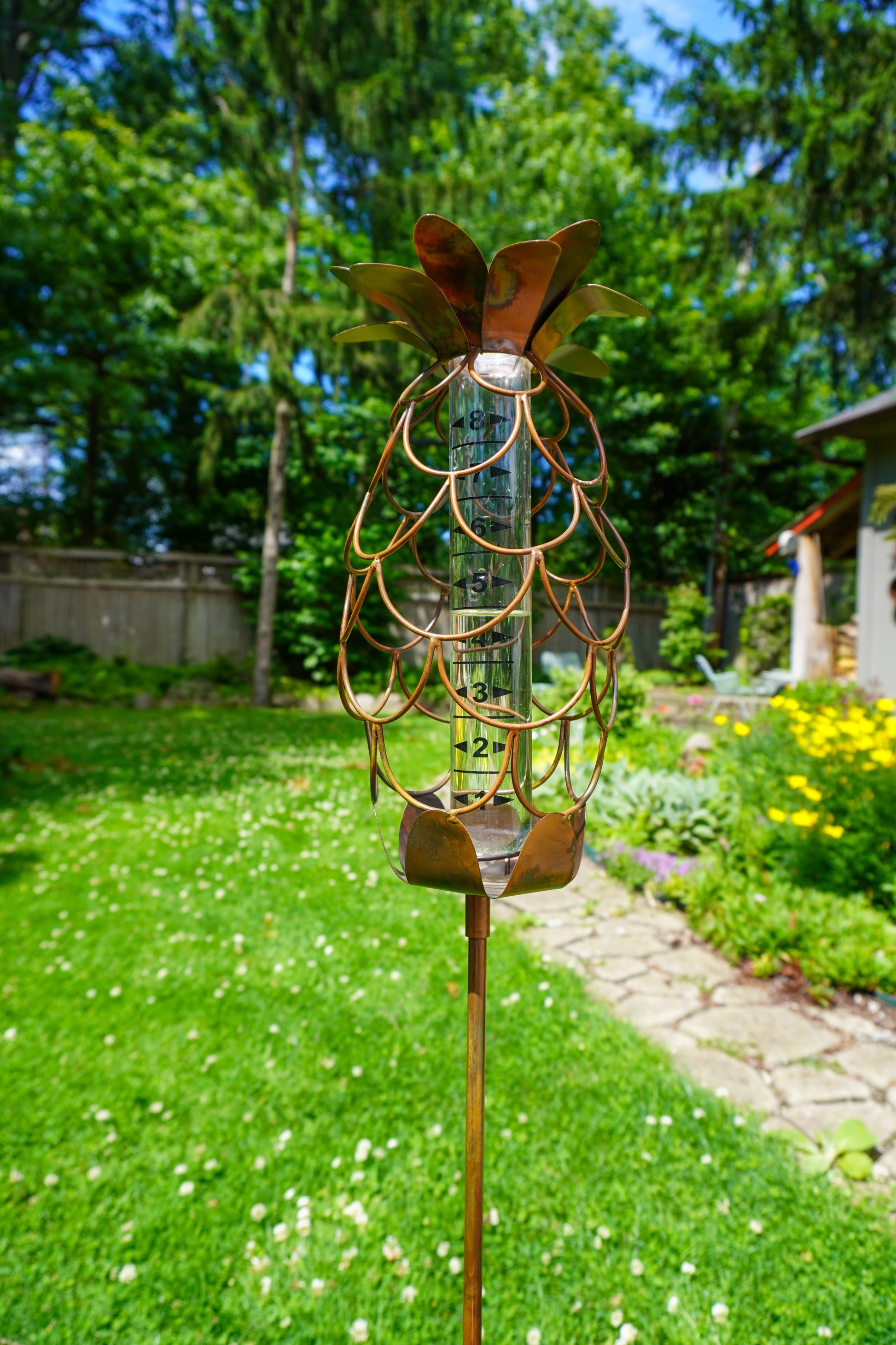 Copper-toned metal pineapple-shaped rain gauge garden stake with glass tube insert, displayed outdoors in a green garden