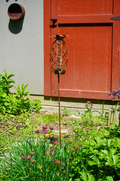 Decorative metal pineapple-shaped rain gauge on a stake displayed in a garden beside a red barn door