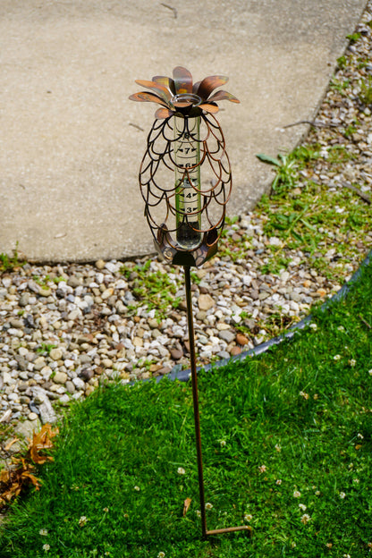 Copper-toned metal pineapple-shaped rain gauge garden stake with glass tube insert, placed in grass near gravel and pavement