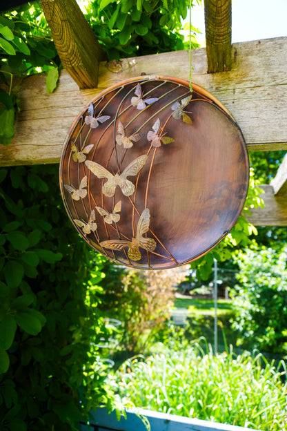 Decorative metal plate with butterfly design hanging on a wooden fence with a garden background