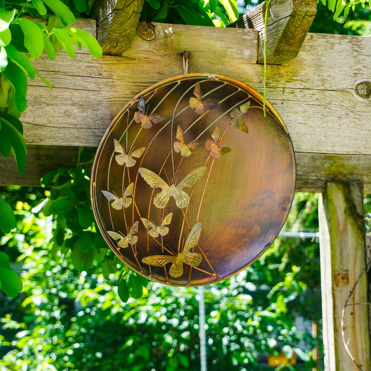 Decorative metal plate with butterfly design hanging on a wooden beam against a green foliage background