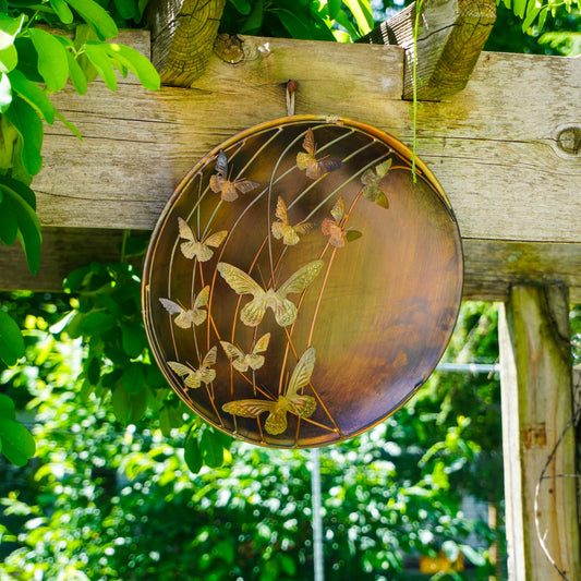 Decorative metal plate with butterfly design hanging on a wooden beam against a green foliage background