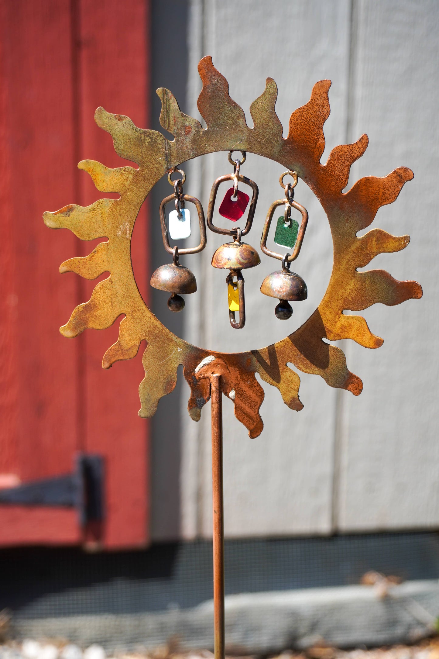 Decorative metal sun-shaped stake with hanging colorful jingle bells against a red and gray background.