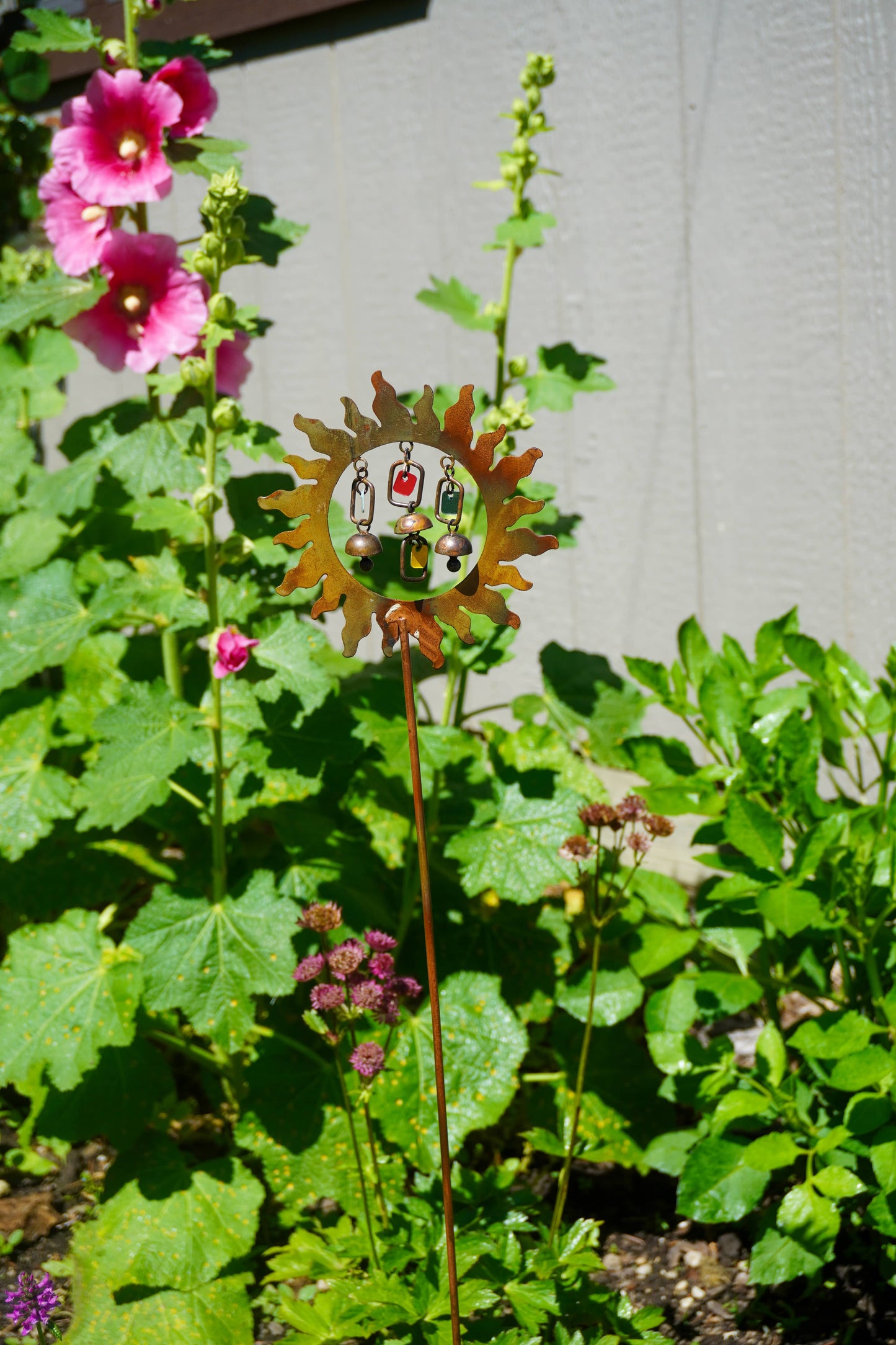 Decorative metal garden stake with sun design in a garden setting with pink flowers and green leaves.