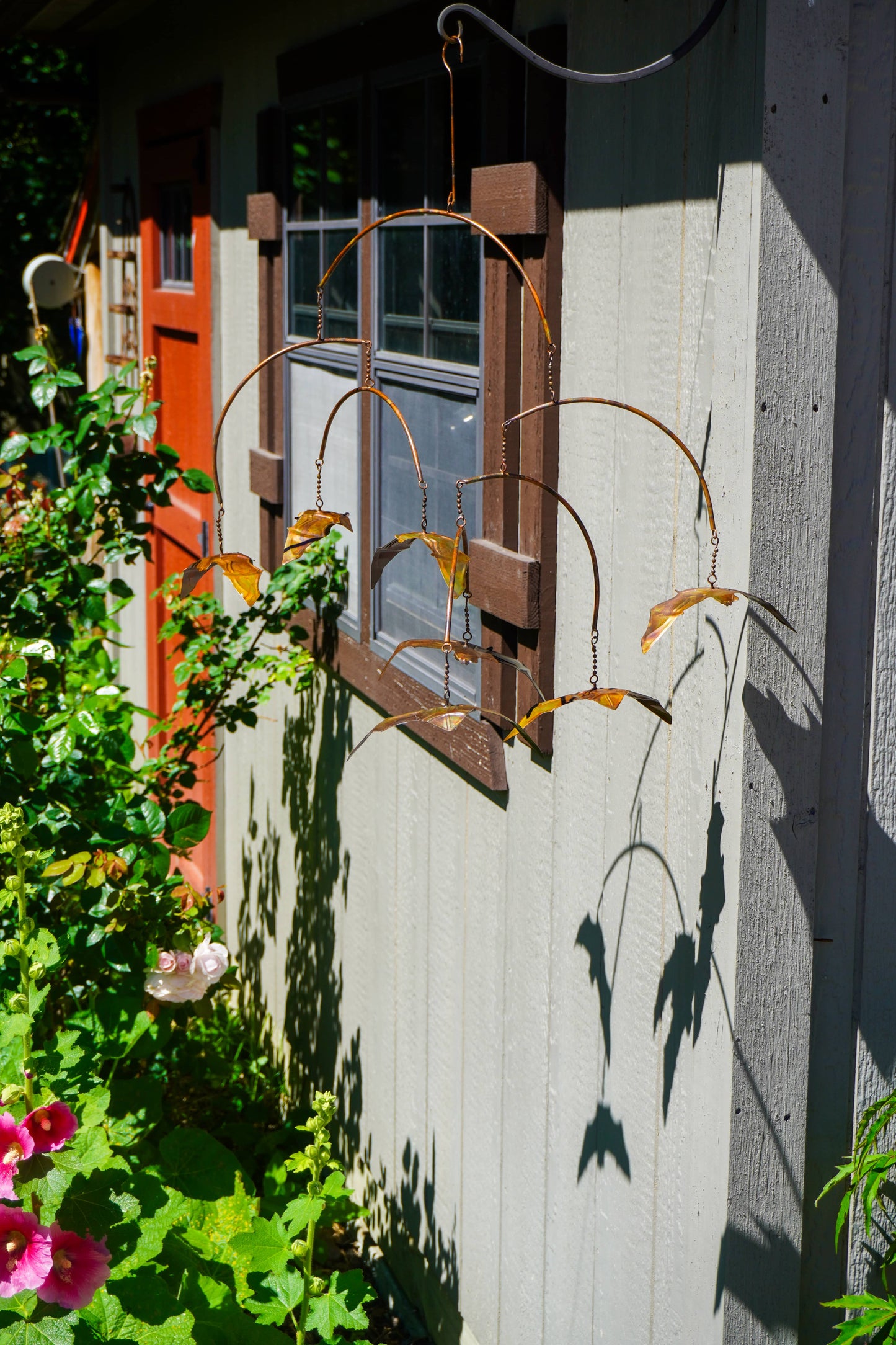 A rusted metal garden wind spinner with bat-like curved blades hangs outdoors against a light gray house wall next to a window and green garden plants.