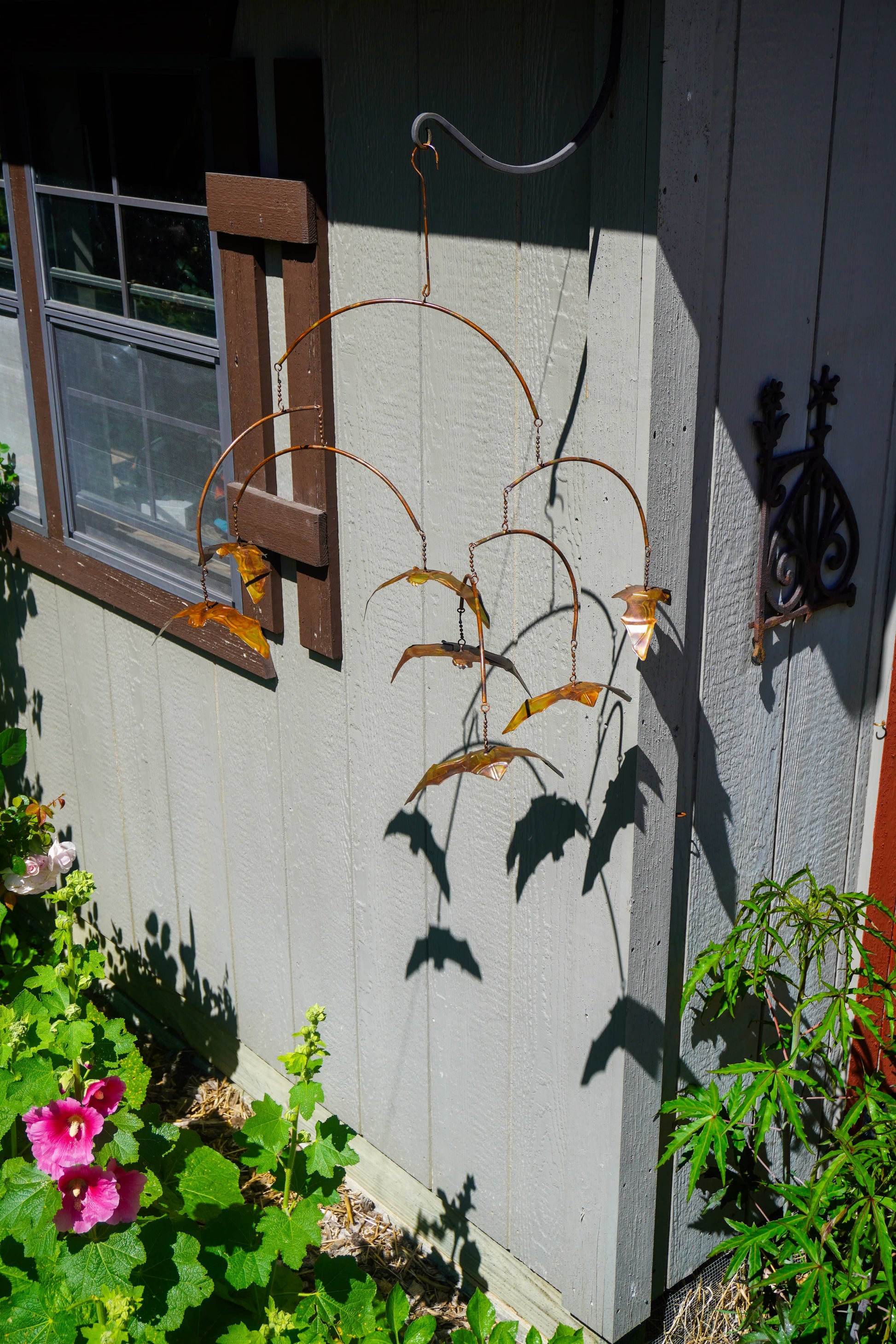 A rustic metal wind spinner with bat-shaped blades hangs on the exterior wall of a house next to a window, casting a distinct shadow on the light gray siding.