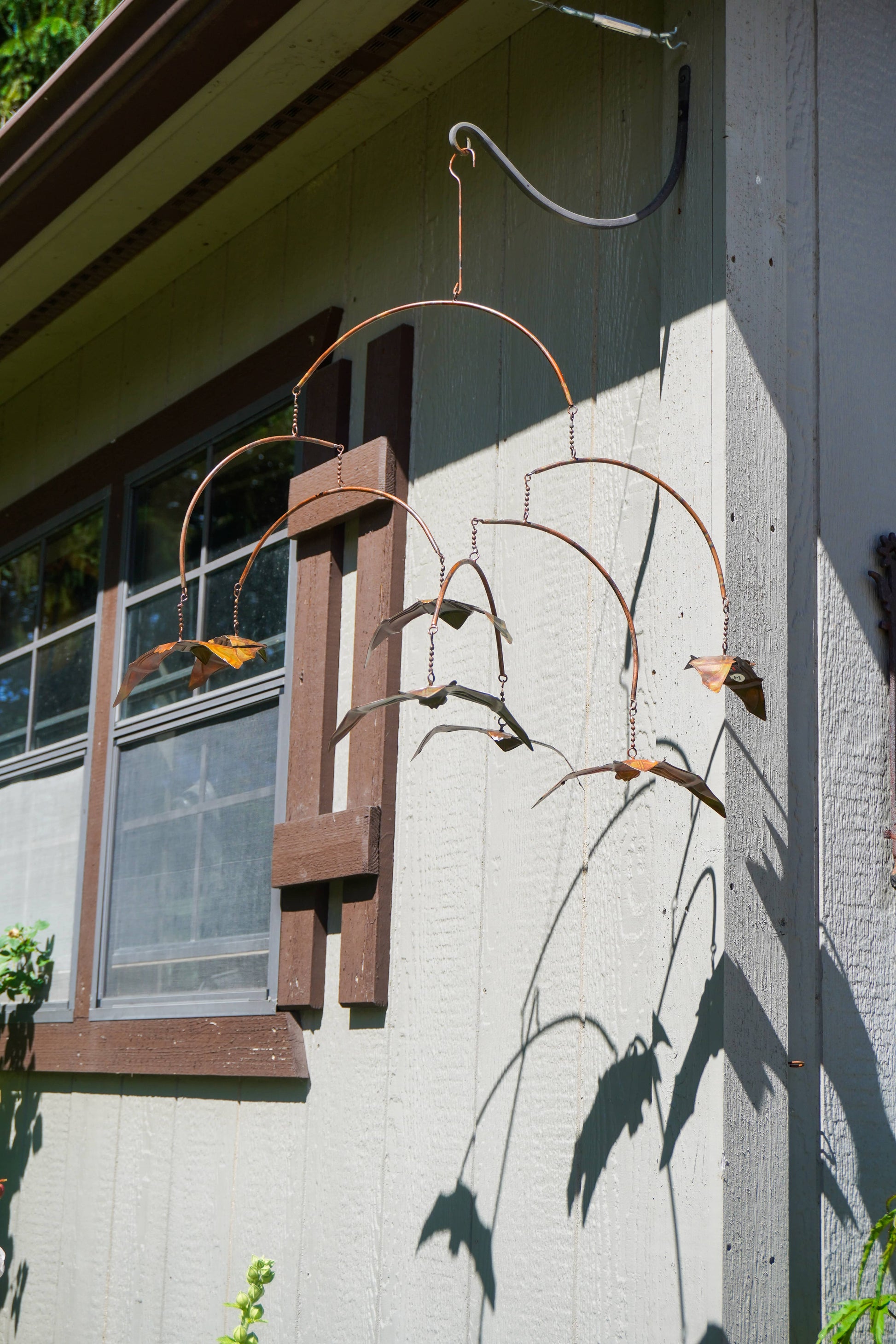 A rusted metal bat mobile hanging from a bracket on the exterior wall of a house, casting a shadow on the stucco wall next to a window.