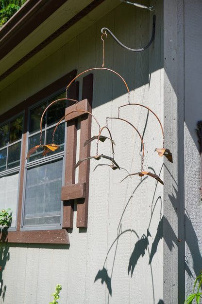 A rusted metal bat mobile hanging from a bracket on the exterior wall of a house, casting a shadow on the stucco wall next to a window.