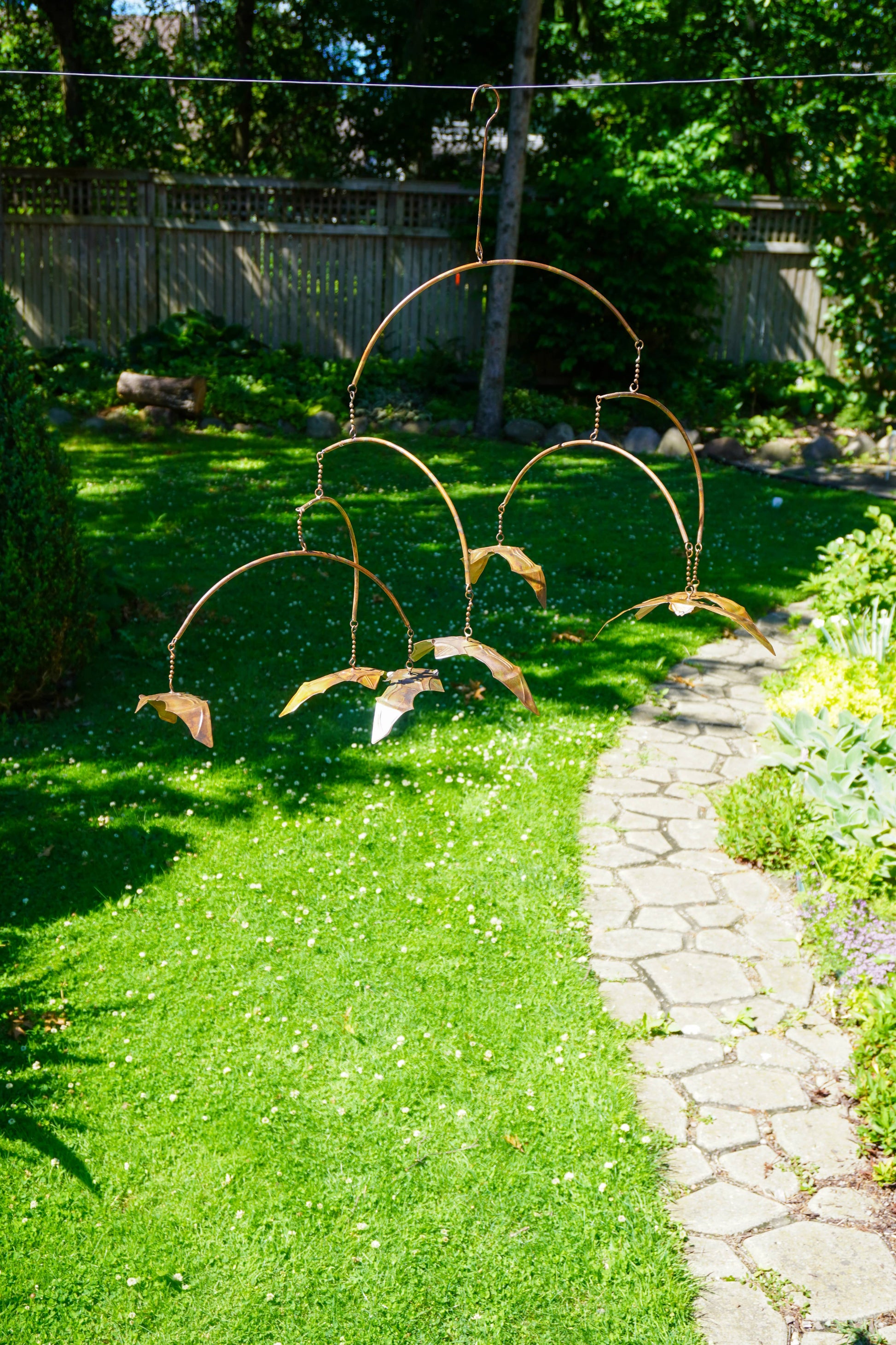 A copper metal garden wind spinner with bat-shaped blades hanging outdoors on a lawn next to a stone pathway.
