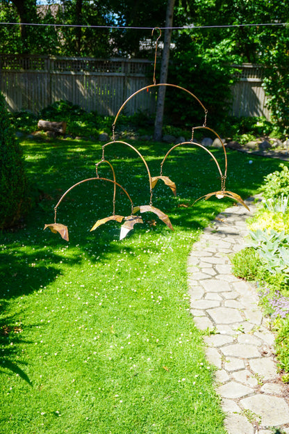 A copper metal garden wind spinner with bat-shaped blades hanging outdoors on a lawn next to a stone pathway.