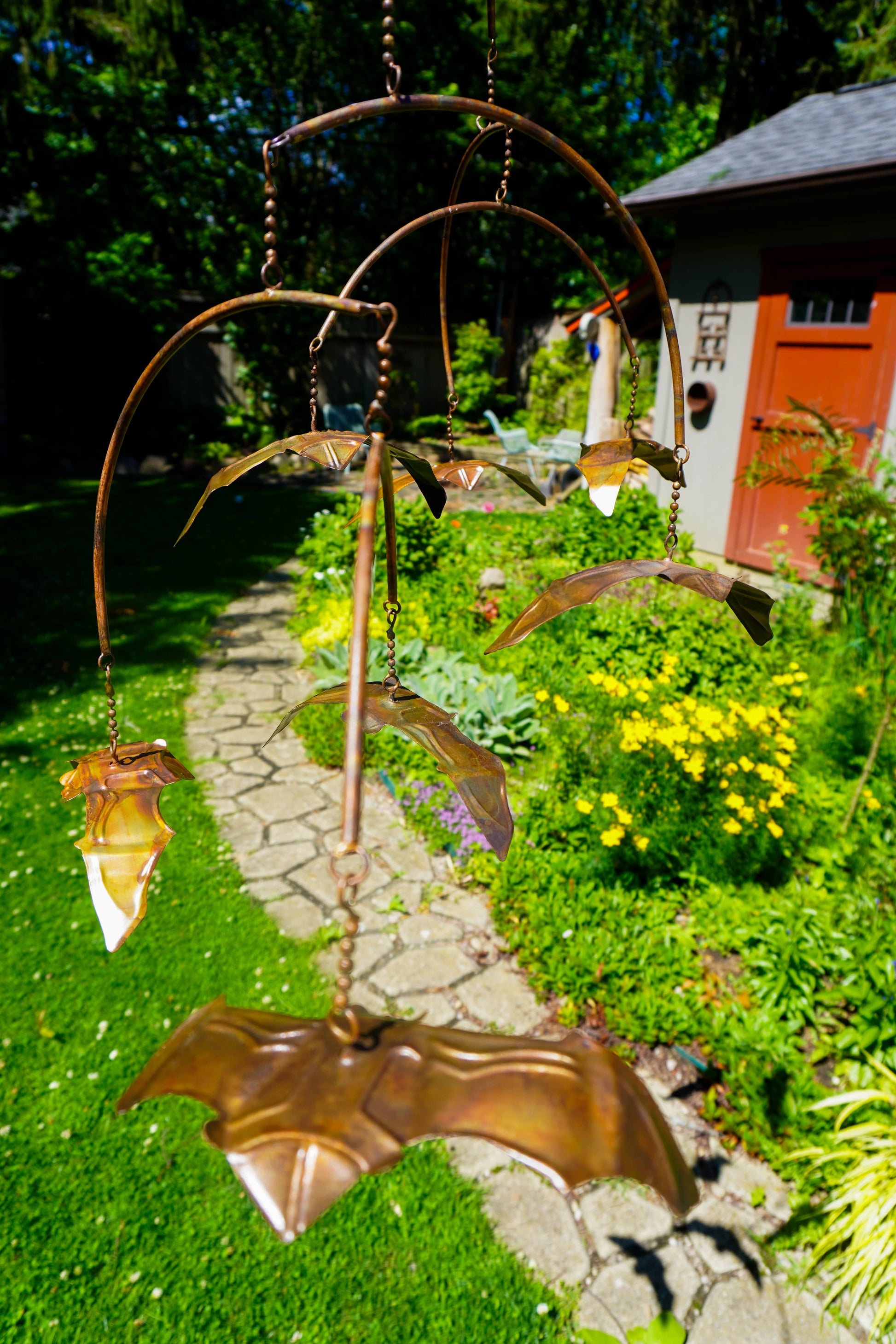 A bronze metal garden mobile with bat-shaped elements and hanging glass shades, photographed outdoors in a sunny garden with a stone path and house in the background.