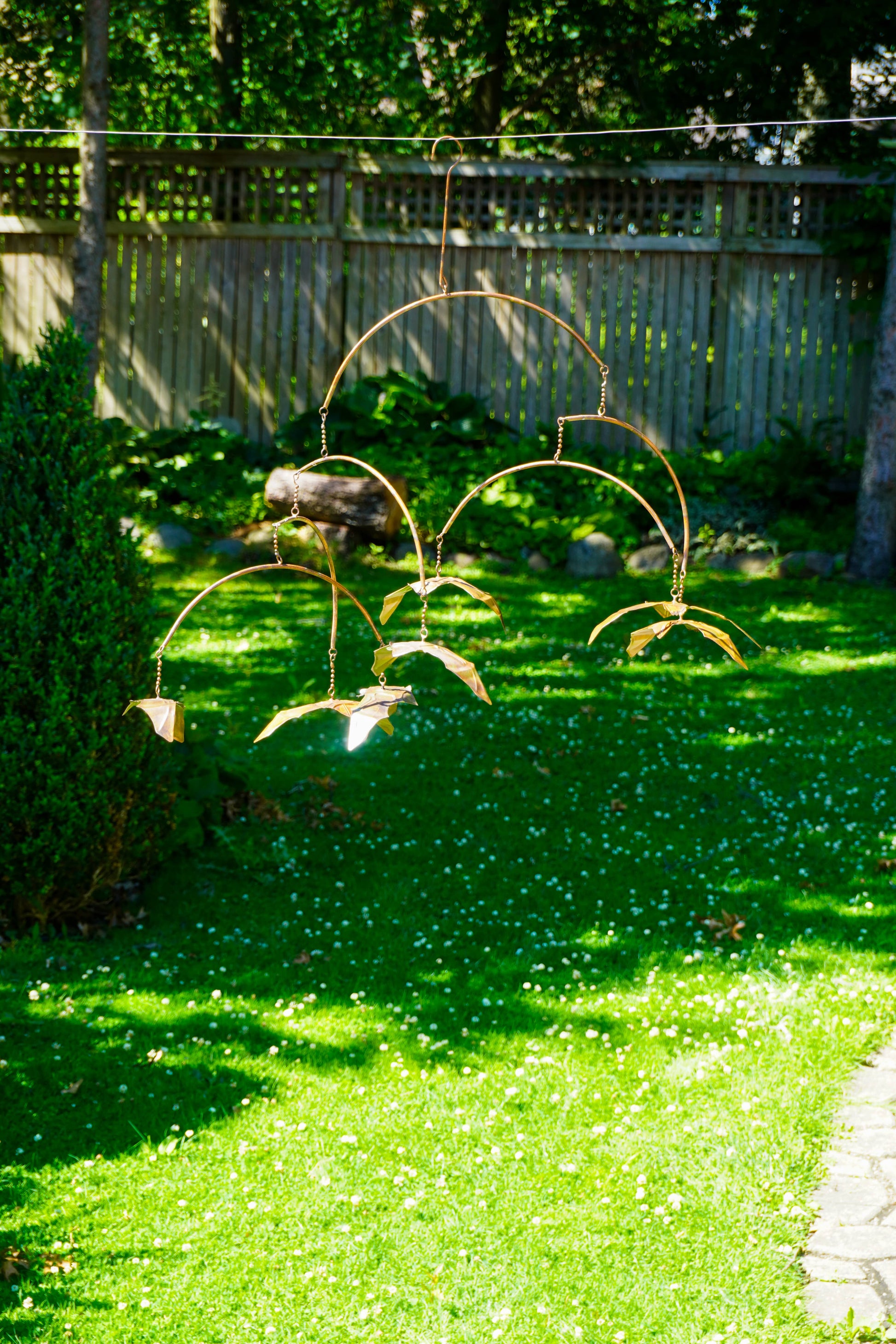 A rustic metal garden wind spinner with bat-like blades hangs outdoors on a grassy lawn in front of a wooden fence.
