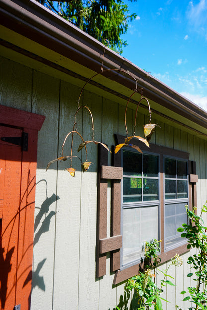 A decorative hanging metal bat wind spinner suspended from a gutter on the exterior of a house with beige siding, a brown window frame, and a red-orange door.
