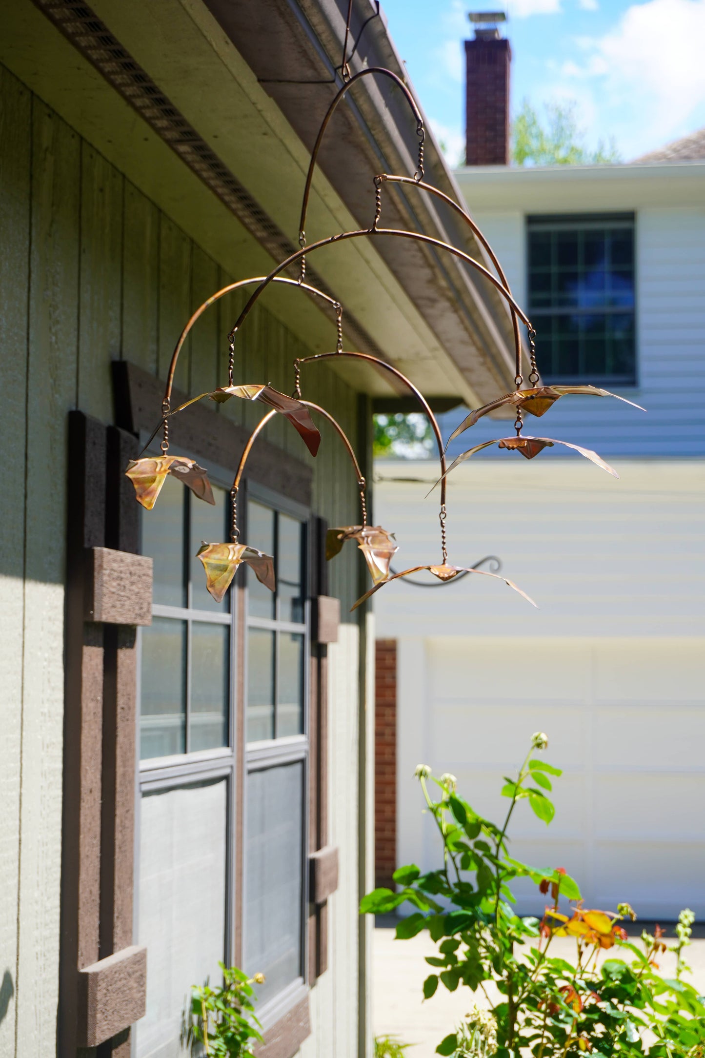 A metal bat wind spinner hanging from a house exterior, with several small birds crafted from metal in flight poses. The wind spinner is shown in a real-world outdoor environment next to a window and house siding.