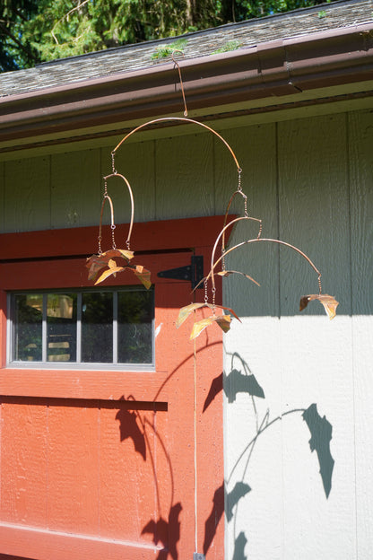 A rust-colored hanging mobile with bat-shaped blades hanging outside a building, casting a shadow on the house wall.