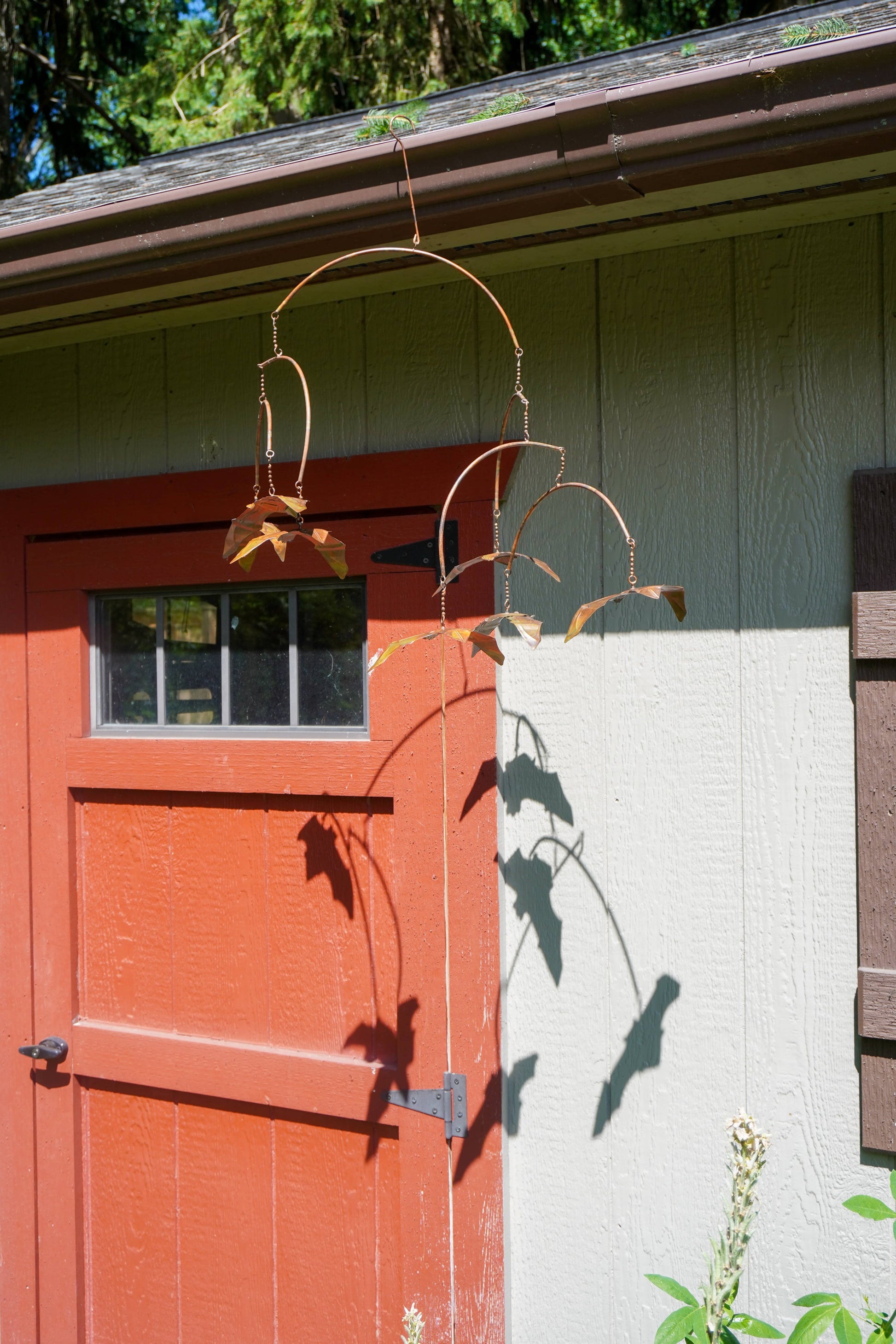 A rustic metal garden mobile with bats hanging in front of a red barn door and light green house siding.