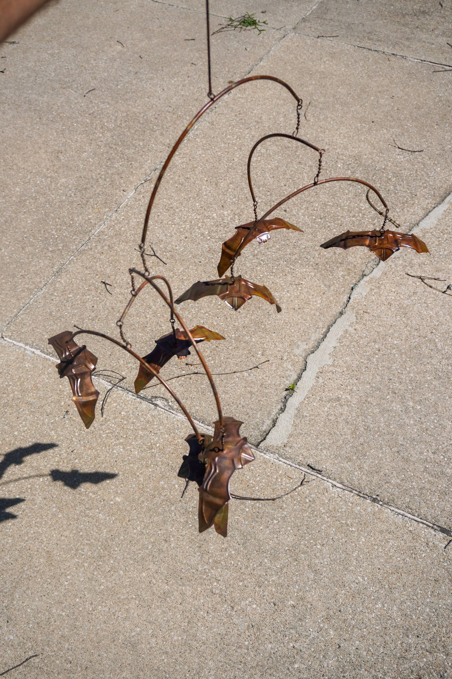 A rusted metal garden wind spinner with curved branches and bat-like shapes, photographed outdoors on a light concrete or stone patio.