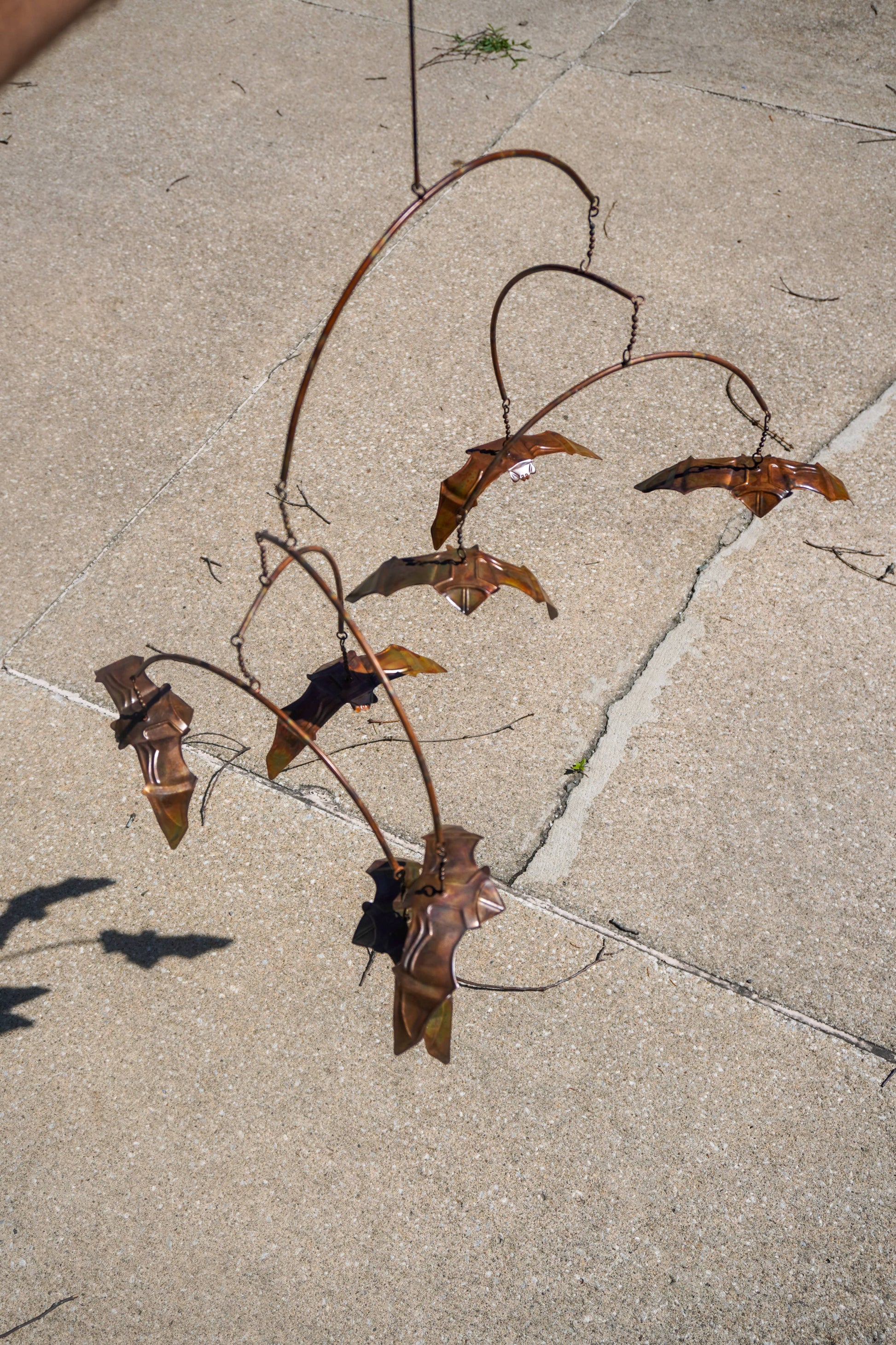 A rusted metal garden wind spinner with curved branches and bat-like shapes, photographed outdoors on a light concrete or stone patio.