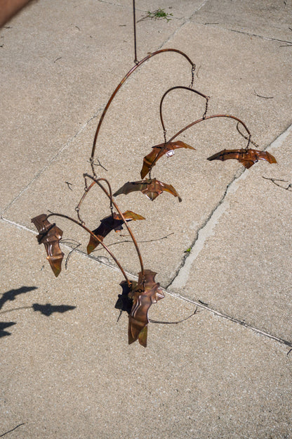 A rusted metal garden wind spinner with curved branches and bat-like shapes, photographed outdoors on a light concrete or stone patio.
