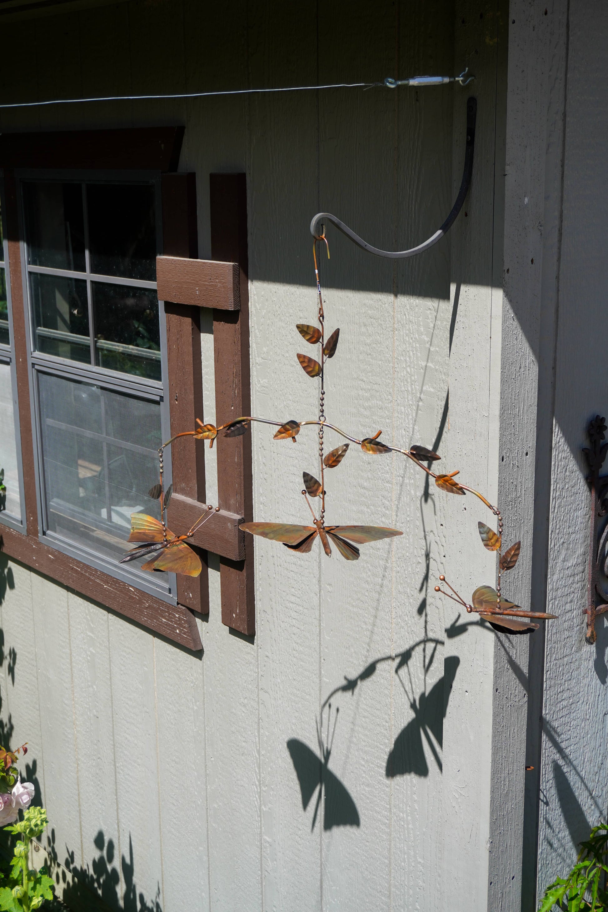 A rustic metal butterfly wind spinner hanging from a wire frame against the exterior wall of a house. The wind spinner features multiple brown and tan butterflies with detailed wings, attached to a thin branch-like structure with small leaves.